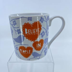 May include: White ceramic mug with a curved handle. The mug features orange heart-shaped designs with the words "BELIEVE IN PEOPLE" and blue floral and bird illustrations. The mug is on a clear surface.