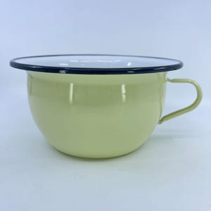 May include: A pale yellow enamelware bowl with a black rim and a handle. The bowl has a rounded shape and a white interior. The handle is curved and attached to the side of the bowl.