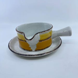 May include: A ceramic gravy boat with a matching saucer. The boat features a white interior and handle, with a yellow and brown striped exterior. The saucer and boat have a speckled, textured appearance. The gravy boat is designed with a spout for easy pouring.