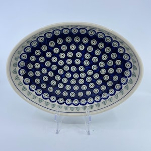 May include: Oval ceramic serving dish with a blue and white pattern. The dish features a dark blue interior with a repeating pattern of white circles with brown centers. The rim is white with a scalloped edge and green accents.