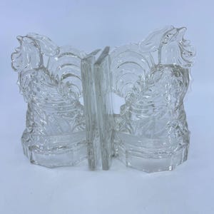 May include: Clear glass bookends with a rooster design. The bookends feature a detailed, textured pattern and a rectangular base. The rooster's head and tail feathers are prominently displayed, adding a decorative touch to any bookshelf.