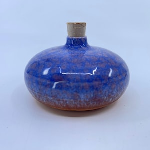 May include: A small, round ceramic bottle with a blue glaze and a cork stopper. The bottle has a speckled pattern with hints of brown and a reddish-brown base. The cork is a light tan color.