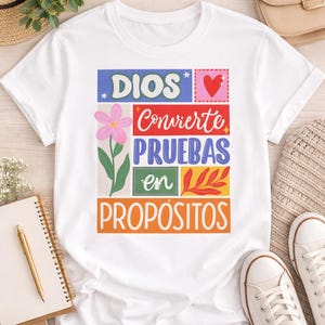 May include: White t-shirt with a colorful graphic design. The design includes the words "DIOS" and "Conviértete PRUEBAS en PROPÓSITOS" in various colors and fonts, along with a pink flower and a red heart. The shirt is paired with white sneakers.