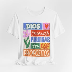 Peut inclure: T-shirt blanc avec un motif graphique coloré. Le motif comprend le mot "DIOS" en bleu, une fleur rose, un cœur rouge et les mots "Convierte PRUEBAS en PROPOSITOS" en plusieurs couleurs. Le t-shirt a des manches courtes.