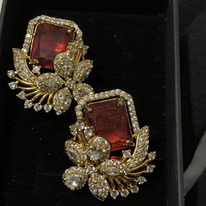 May include: A pair of gold-toned earrings with a square, deep red gemstone center, surrounded by a halo of small, clear crystals. The earrings feature a floral design with additional clear crystals and gold-toned accents.