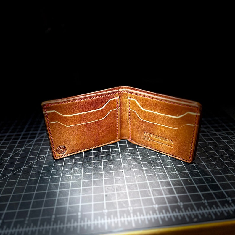 以下が含まれることがあります： 赤いステッチのディテールと前面に「HANDMADE LEATHER CO.」の刻印が施された茶色の革財布です。