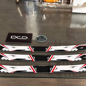 Può includere: Tre finiture decorative per auto nere, bianche e rosse con un design geometrico. Sono presenti anche una scheda nera con la scritta "DCD PAINTWOOD" e un adesivo argentato "LID MODS". Pezzi per la personalizzazione del veicolo.