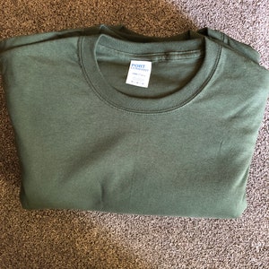 May include: A folded green long-sleeved t-shirt with a white tag that reads "PORT COMPANY" and "COMFORT COLORS" with the size "M" printed on it.
