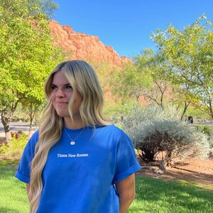May include: A woman wearing a light blue t-shirt with the text "Times New Roman" embroidered on the front. She is standing in front of a red rock formation and green grass.