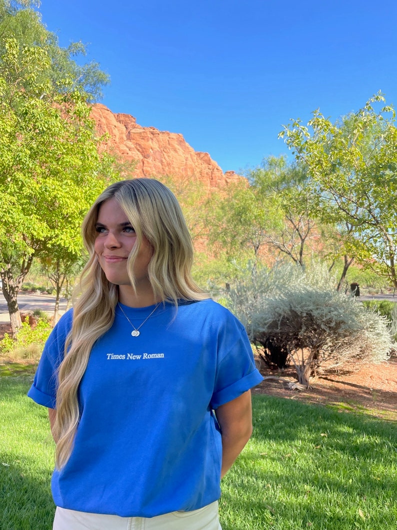 May include: A woman wearing a light blue t-shirt with the text "Times New Roman" embroidered on the front. She is standing in front of a red rock formation and green grass.