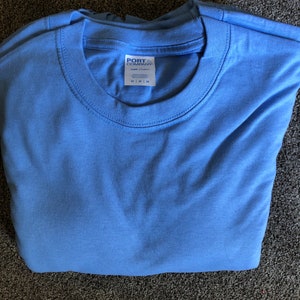 May include: A folded light blue t-shirt with a white tag that reads "PORT COMPANY" and "100% Cotton" with size information.