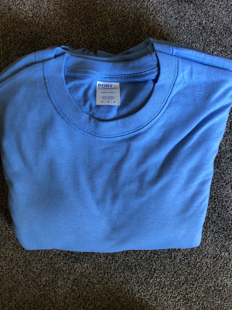 May include: A folded light blue t-shirt with a white tag that reads "PORT COMPANY" and "100% Cotton" with size information.