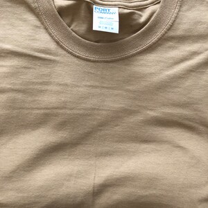May include: A folded tan t-shirt with a white tag that reads "PORT COMPANY" and "One Cotton" with size options "S | M | L"