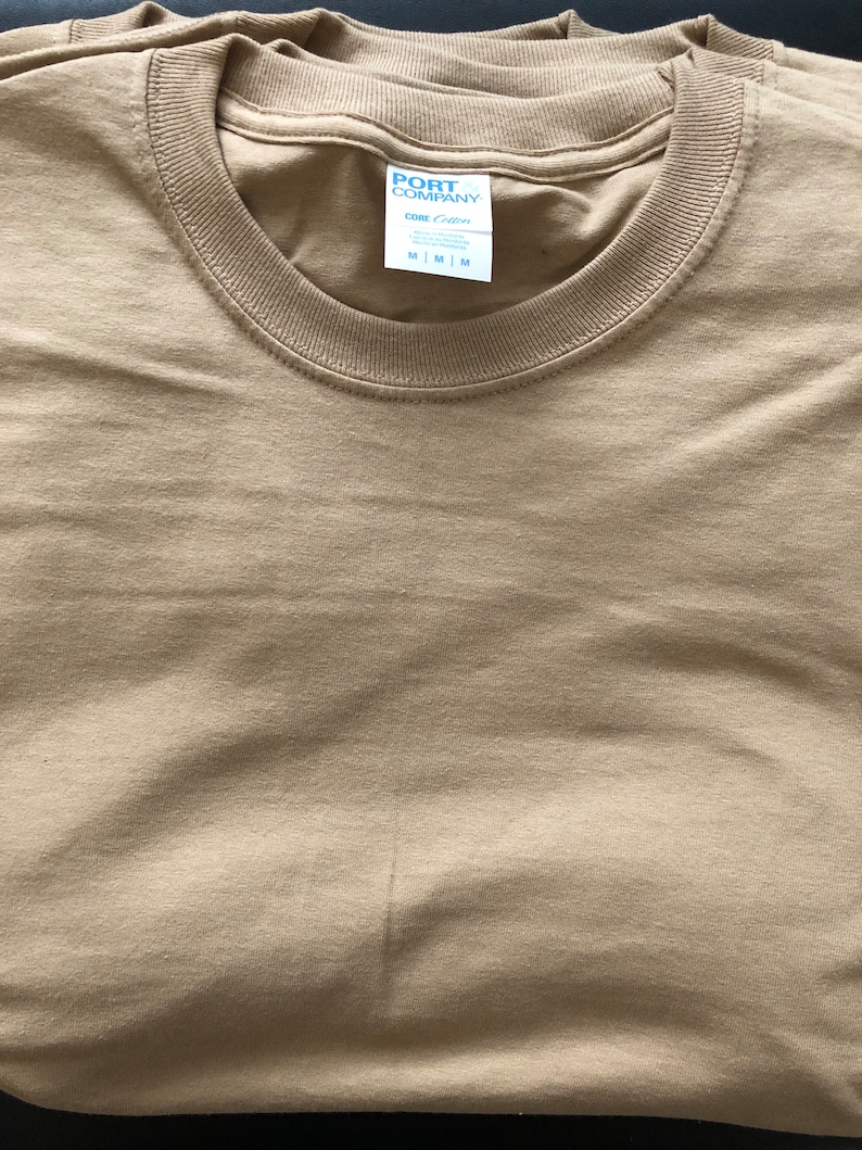May include: A folded tan t-shirt with a white tag that reads "PORT COMPANY" and "One Cotton" with size options "S | M | L"