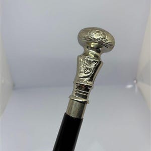 May include: Silver-colored walking cane with a black handle. The top of the cane features a decorative floral design and a detailed silver-colored metalwork. The handle is a smooth, polished black.