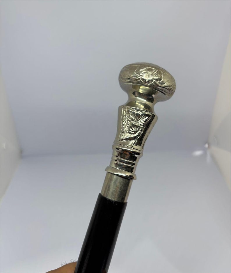 May include: Silver-colored walking cane with a black handle. The top of the cane features a decorative floral design and a detailed silver-colored metalwork. The handle is a smooth, polished black.