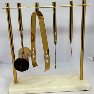 Pode incluir: Um conjunto de ferramentas de bar douradas com uma base de mármore. O conjunto inclui um jigger, pinças, um pilão, uma colher de bar e um coador. As ferramentas são suspensas de uma barra horizontal.