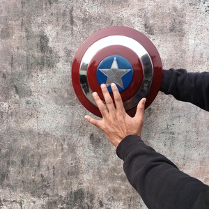 May include: A Captain America shield in red, white, and blue, featuring a silver star emblem. The shield has a circular design with a metallic silver ring. The shield is held against a textured grey wall.