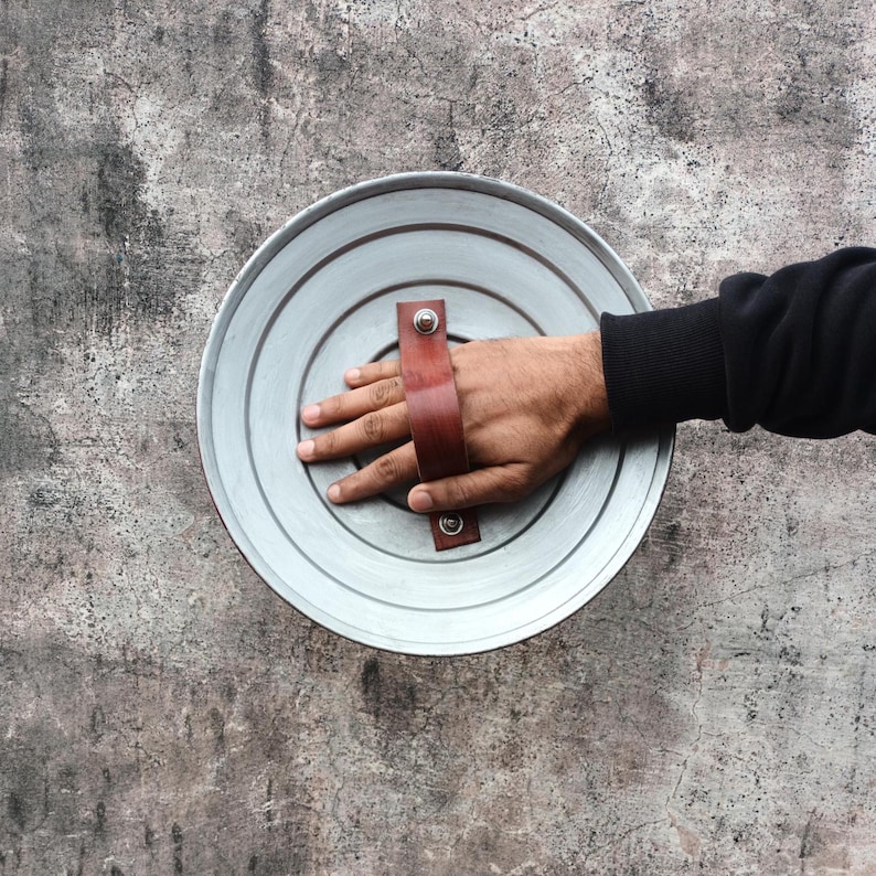 May include: A circular, silver-toned metal shield featuring concentric circles. It has a brown leather handle secured with rivets. The shield is held by a hand, with a dark sleeve visible.