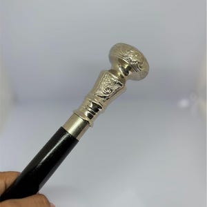 Puede incluir: Bastón plateado con un mango de madera negra. El mango es adornado con diseños florales grabados. El bastón se sostiene en ángulo, mostrando los detalles del mango y la madera negra pulida.