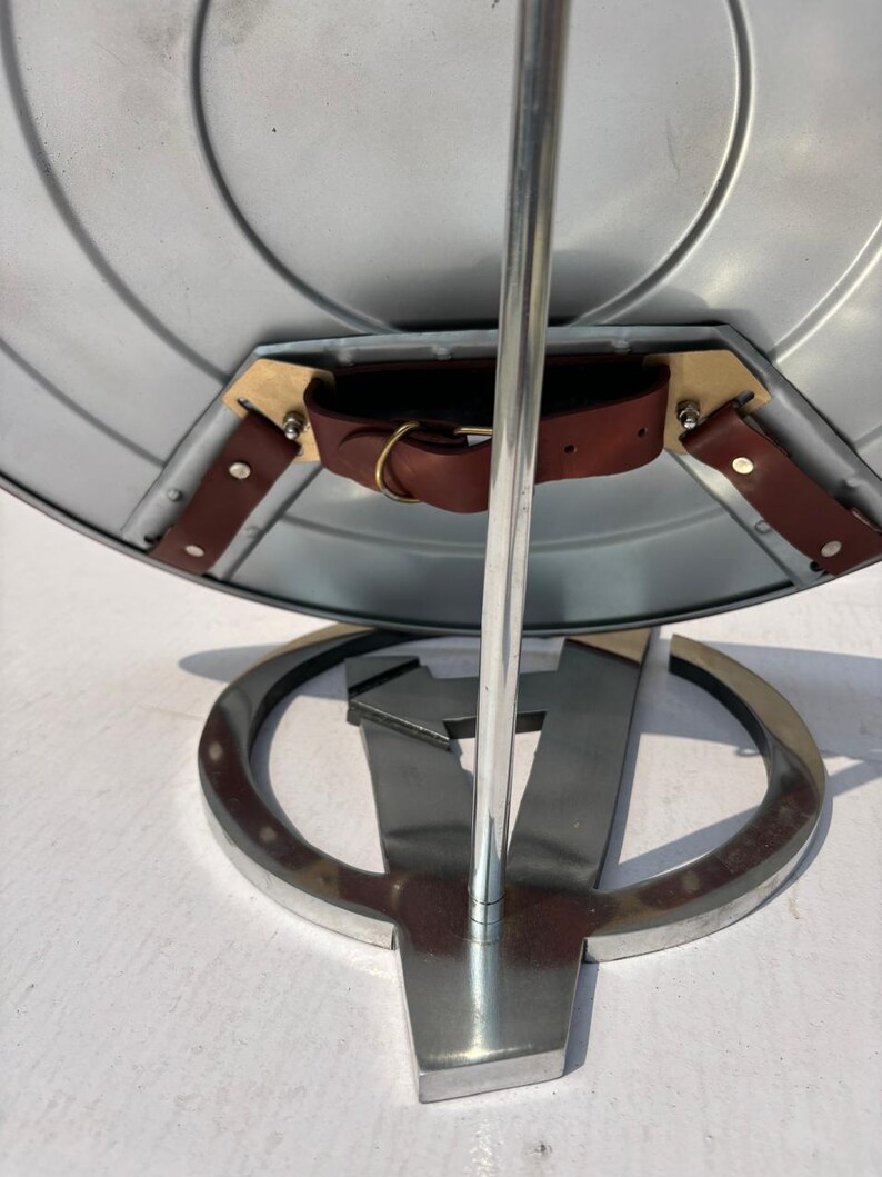 K&ouml;nnte beinhalten: Ein silberner Schild mit einem braunen Lederriemen und Messingbeschl&auml;gen wird auf einem Metallst&auml;nder pr&auml;sentiert. Der St&auml;nder hat eine silberne Stange und einen Sockel mit einem stilisierten "V"-Design. Der Schild hat konzentrische kreisf&ouml;rmige Rillen.