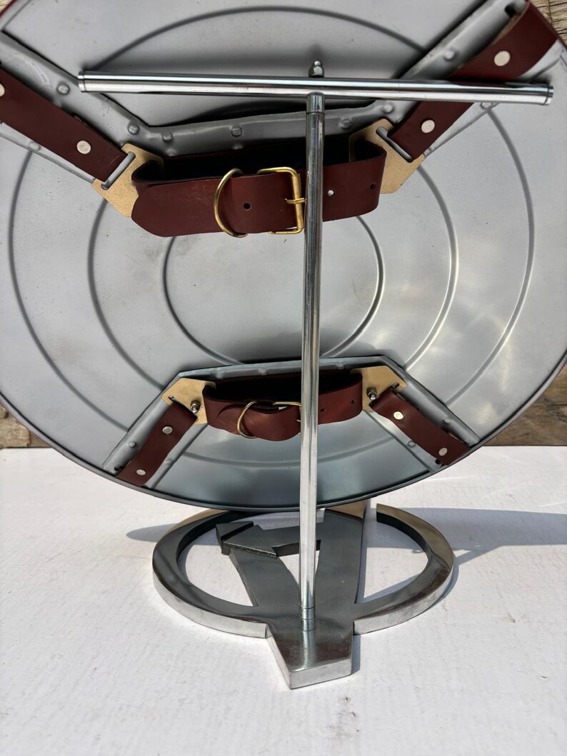 K&ouml;nnte beinhalten: Ein silberner Schild mit braunen Lederriemen und goldenen Schnallen, ausgestellt auf einem Metallst&auml;nder. Der St&auml;nder zeigt das Avengers-Logo. Der Schild hat ein kreisf&ouml;rmiges Design.