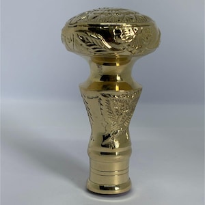 May include: A gold-colored, ornate walking cane handle. The handle features detailed floral and leaf patterns, with a rounded top and a cylindrical base. The metal has a polished, reflective surface, and the design is symmetrical.