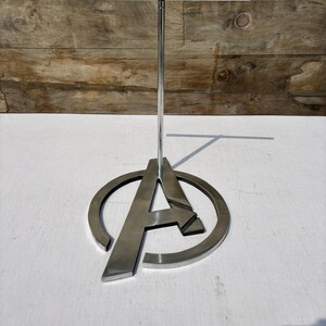 K&ouml;nnte beinhalten: Ein silberner Metall-Displayst&auml;nder mit dem Avengers "A"-Logo. Der St&auml;nder hat eine horizontale Stange oben und eine kreisf&ouml;rmige Basis mit dem Buchstaben "A" in der Mitte. Der St&auml;nder ist aus gl&auml;nzendem Metall gefertigt und eignet sich zur Warenpr&auml;sentation.