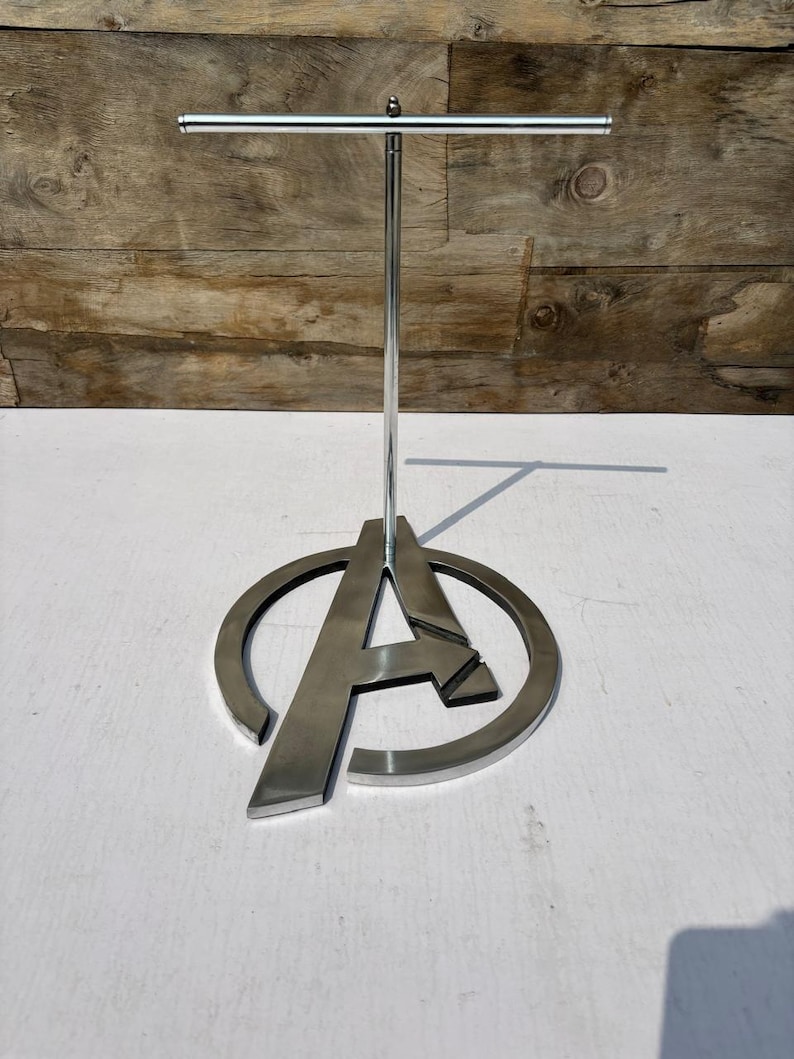 K&ouml;nnte beinhalten: Ein silberner Metall-Displayst&auml;nder mit dem Avengers "A"-Logo. Der St&auml;nder hat eine horizontale Stange oben und eine kreisf&ouml;rmige Basis mit dem Buchstaben "A" in der Mitte. Der St&auml;nder ist aus gl&auml;nzendem Metall gefertigt und eignet sich zur Warenpr&auml;sentation.