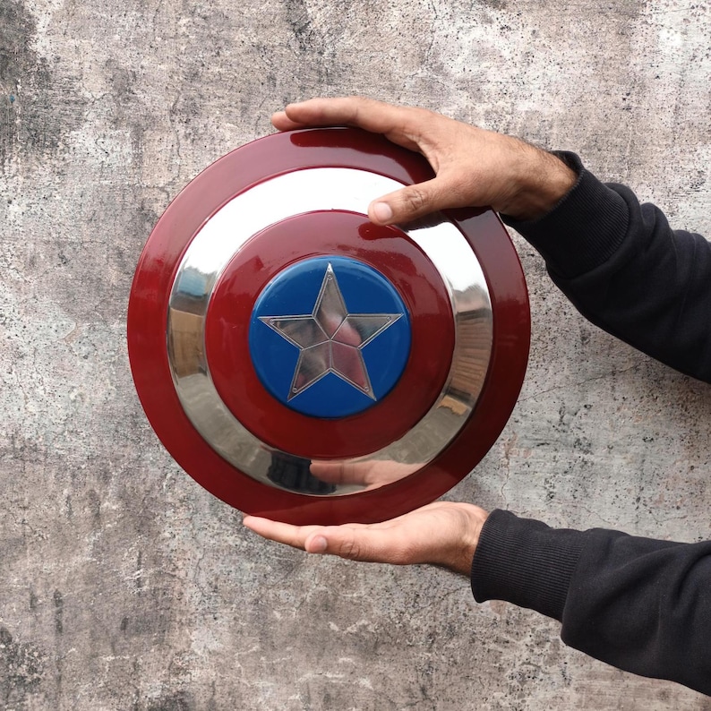 May include: A circular shield in red, white, and blue, featuring a central star emblem. The design includes a silver band separating the red and blue sections. The shield is held by two hands.
