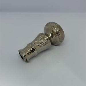 May include: A silver-colored bottle stopper with an ornate design. The stopper features a detailed floral pattern on the top and an eagle emblem on the body. The metal has a polished finish, reflecting light.