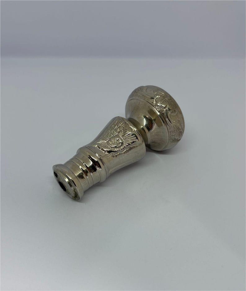 May include: A silver-colored bottle stopper with an ornate design. The stopper features a detailed floral pattern on the top and an eagle emblem on the body. The metal has a polished finish, reflecting light.