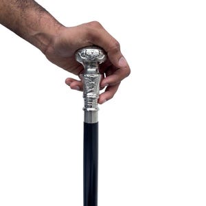 May include: A decorative walking cane with a silver-colored handle and a dark blue shaft. The handle features an ornate floral design. The cane is held by a hand against a white background.