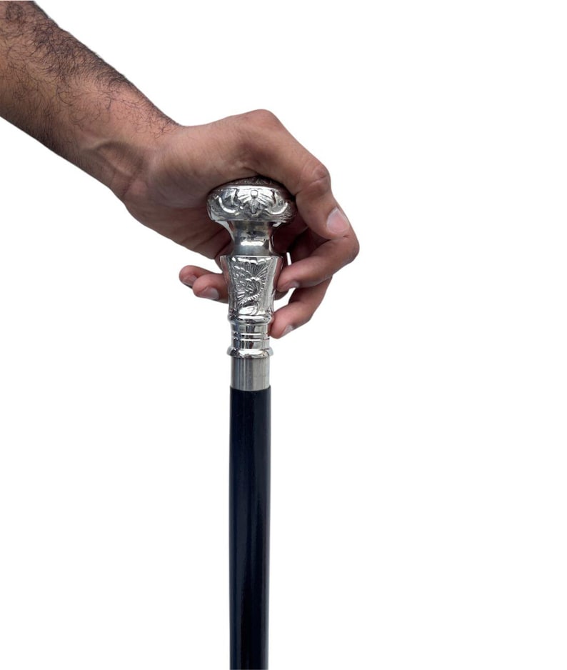 May include: A decorative walking cane with a silver-colored handle and a dark blue shaft. The handle features an ornate floral design. The cane is held by a hand against a white background.