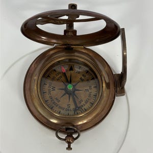 Puede incluir: Una brújula de latón vintage con tapa con bisagras y un reloj de sol. La esfera de la brújula presenta marcas direccionales y una aguja que apunta hacia el norte. La brújula es una herramienta de navegación, y el reloj de sol es un dispositivo para medir el tiempo.