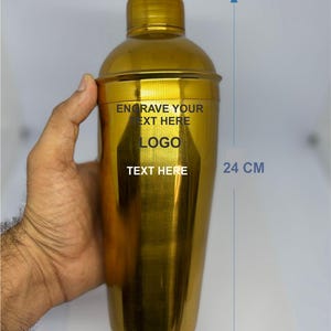 May include: A gold cocktail shaker, approximately 24 cm tall, held by a hand. The shaker has a cap and a cylindrical body. Text on the shaker reads "ENGRAVE YOUR TEXT HERE", "LOGO", and "TEXT HERE".