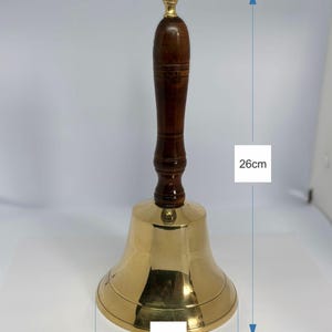 Op de afbeelding: Een messing bel met een donkerbruine houten handgreep en een goudkleurige bovenkant. De bel is 12 cm breed en 26 cm hoog. De bel heeft een klassiek ontwerp en is geschikt voor diverse doeleinden.