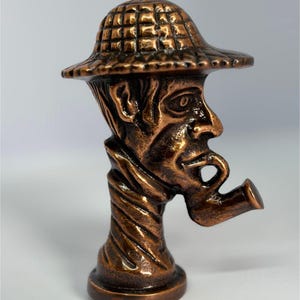 Può includere: Un tappo per bottiglia decorativo color bronzo raffigurante il volto scolpito di un uomo con un cappello e che fuma la pipa. Il tappo ha un design dettagliato e strutturato e una base rotonda.