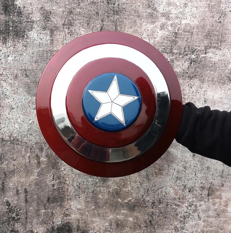 May include: A circular shield in red, white, and blue, with a silver border and a white star on a blue central disc. The shield is held by a person wearing a black long-sleeve shirt.