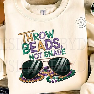 May include: A white sweatshirt with the text "Throw Beads, Not Shade" in green, purple, and gold lettering. A pair of sunglasses with green, purple, and gold beads are on top of the text.