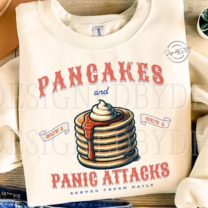May include: A beige sweatshirt with a graphic design of a stack of pancakes with syrup and whipped cream. The text reads "Pancakes and Panic Attacks Served Fresh Daily" with a buy one get one free offer.