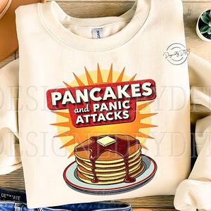 Puede incluir: Una sudadera de color crema con un gráfico de estilo vintage de una pila de panqueques con jarabe y mantequilla. El gráfico dice "Pancakes and Panic Attacks".