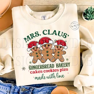 K&ouml;nnte beinhalten: Ein cremefarbener Pullover mit dem Text "MRS. CLAUS, GINGERBREAD BAKERY cakes cookies pies made with love" und drei Lebkuchenm&auml;nnern mit roten H&uuml;ten.