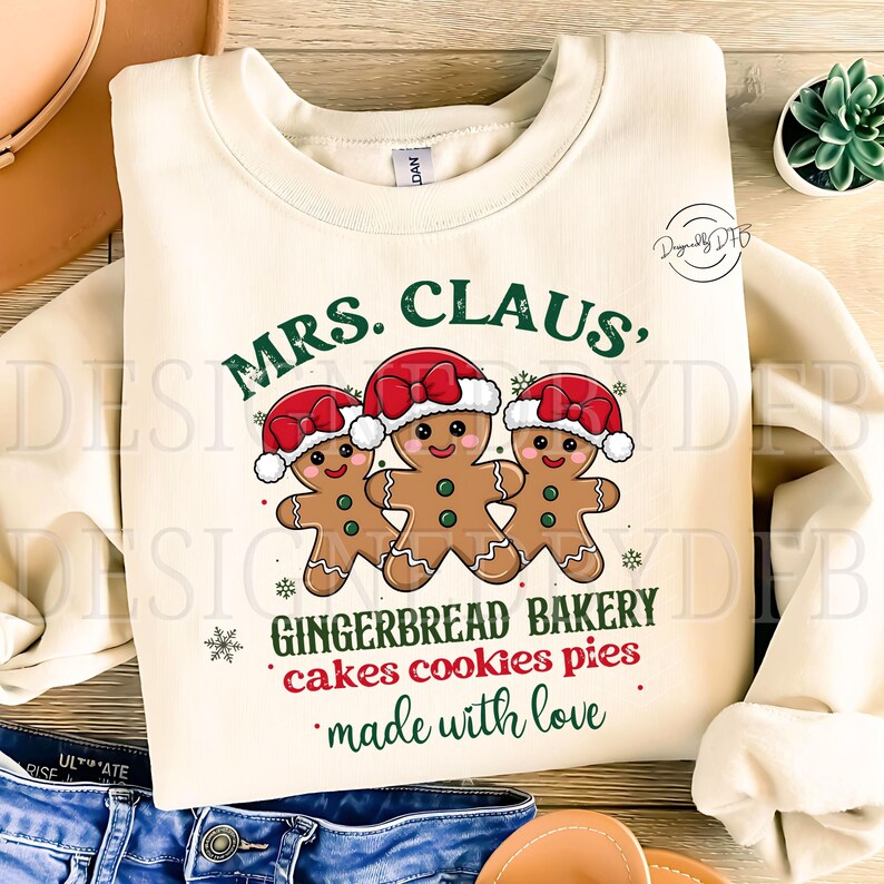 K&ouml;nnte beinhalten: Ein cremefarbener Pullover mit dem Text "MRS. CLAUS, GINGERBREAD BAKERY cakes cookies pies made with love" und drei Lebkuchenm&auml;nnern mit roten H&uuml;ten.