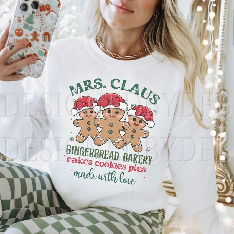 K&ouml;nnte beinhalten: Wei&szlig;es Langarmshirt mit dem Text "MRS. CLAUS GINGERBREAD BAKERY cakes cookies pies made with love."  Drei Lebkuchenm&auml;nner sind in der Mitte des Shirts abgebildet.