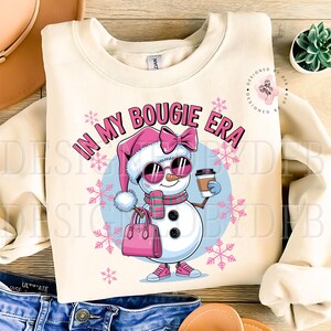 May include: A cream-colored sweatshirt with a snowman wearing sunglasses, a pink hat, and a pink bow. The snowman is holding a cup of coffee and a pink purse. The text "In My Bougie Era" is printed on the sweatshirt.
