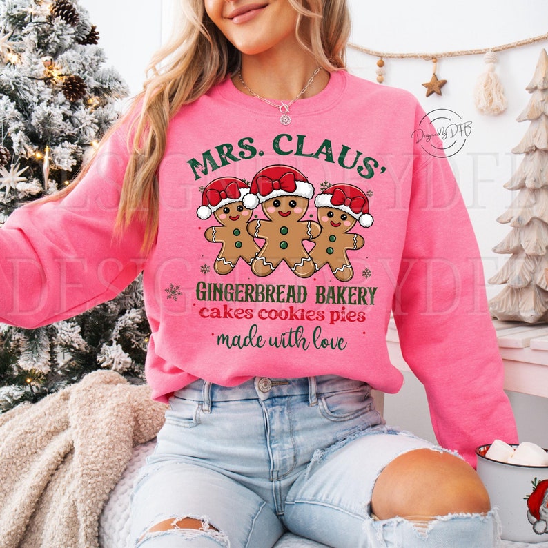 K&ouml;nnte beinhalten: Rosafarbener Pullover mit dem Text "MRS. CLAUS' GINGERBREAD BAKERY cakes cookies pies made with love" und drei Lebkuchenm&auml;nnern mit Weihnachtsm&uuml;tzen.