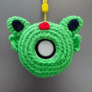 May include: A green crocheted case for an electronic device, shaped like a frog. The case has dark blue accents on the ears and a red dot. A yellow lanyard is attached to the top.