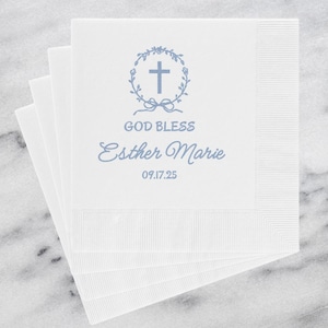 May include: White paper napkins with a blue floral cross design, the words "GOD BLESS" and the name "Esther Marie" are printed on the napkins. The date "09.17.25" is also printed on the napkins. The napkins are stacked on a white marble surface.