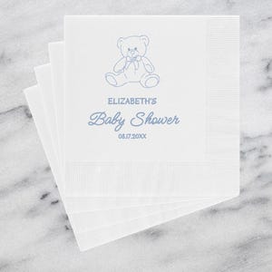 May include: White paper napkins with a blue teddy bear illustration and the text "Elizabeth's Baby Shower" and the date 08.17.20XX. The napkins are stacked on a white and gray marble surface.